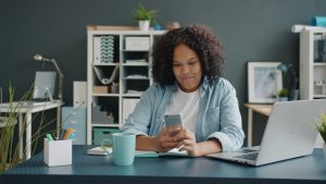Woman using smartphone and laptop in modern office workspace for daily productivity tasks. Photo by Unsplash.